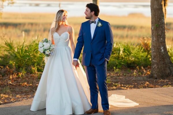 A bride in a white gown holding a bouquet stands beside a groom in a blue suit. They’re outdoors, holding hands, smiling at each other, with greenery and water in the background.