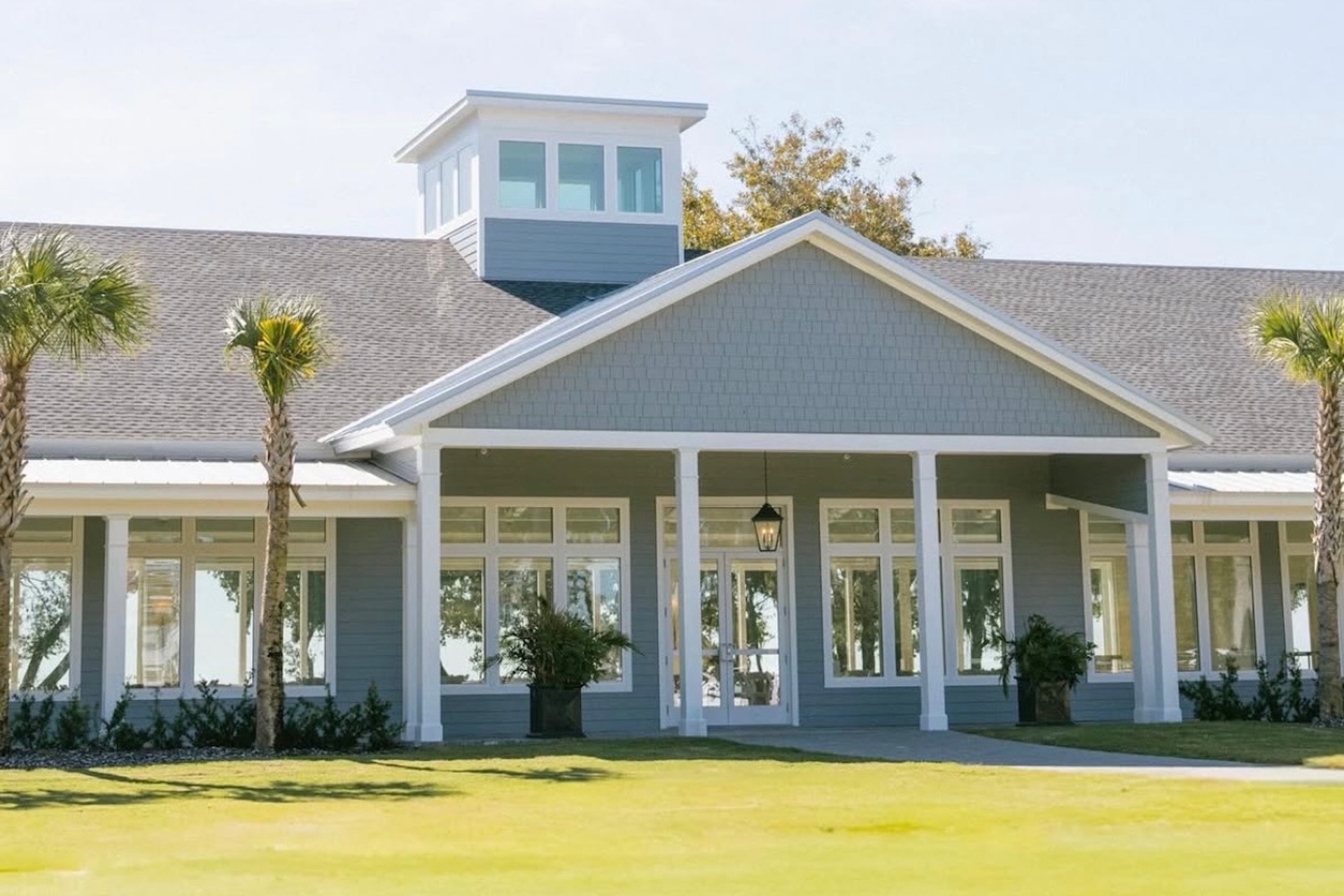 A modern, light gray building with white trim, large windows, and a covered entrance, surrounded by palm trees and green grass under a clear blue sky.