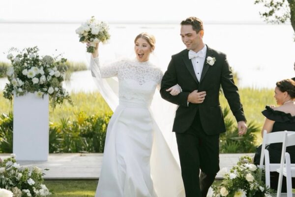 A joyful bride in a white gown holds a bouquet up while walking arm-in-arm with the smiling groom in a black tuxedo outdoors, surrounded by greenery and floral arrangements.