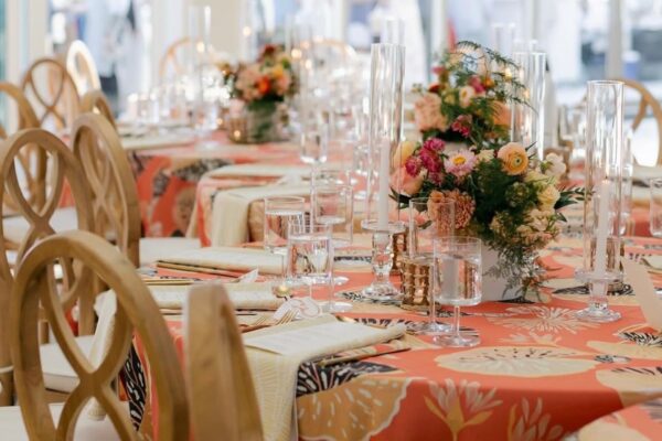 A close-up of a decorated banquet table featuring floral centerpieces, tall glass candleholders, glassware, beige napkins, and wooden chairs. The tablecloth is orange with a tropical leaf pattern.