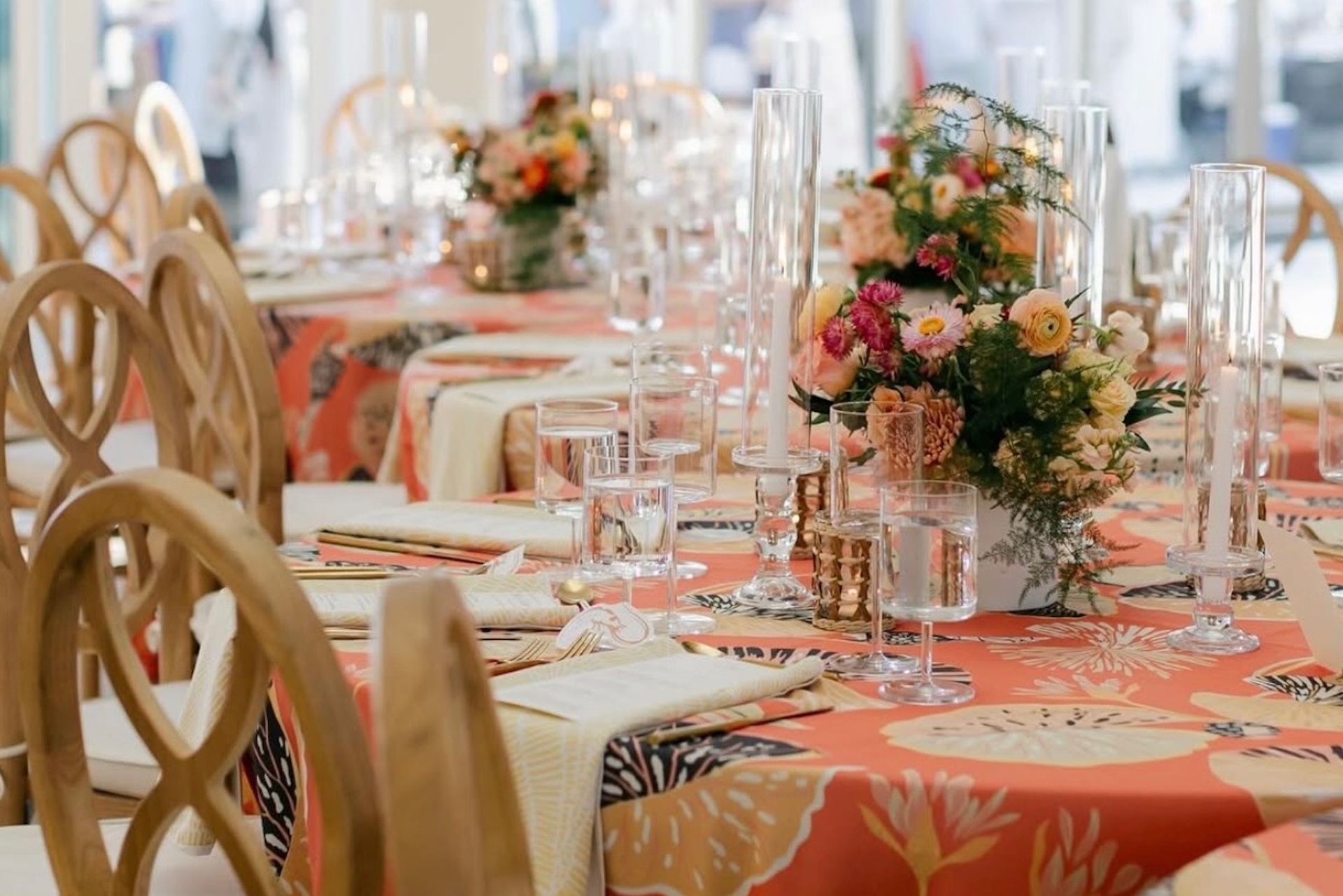 A close-up of a decorated banquet table featuring floral centerpieces, tall glass candleholders, glassware, beige napkins, and wooden chairs. The tablecloth is orange with a tropical leaf pattern.