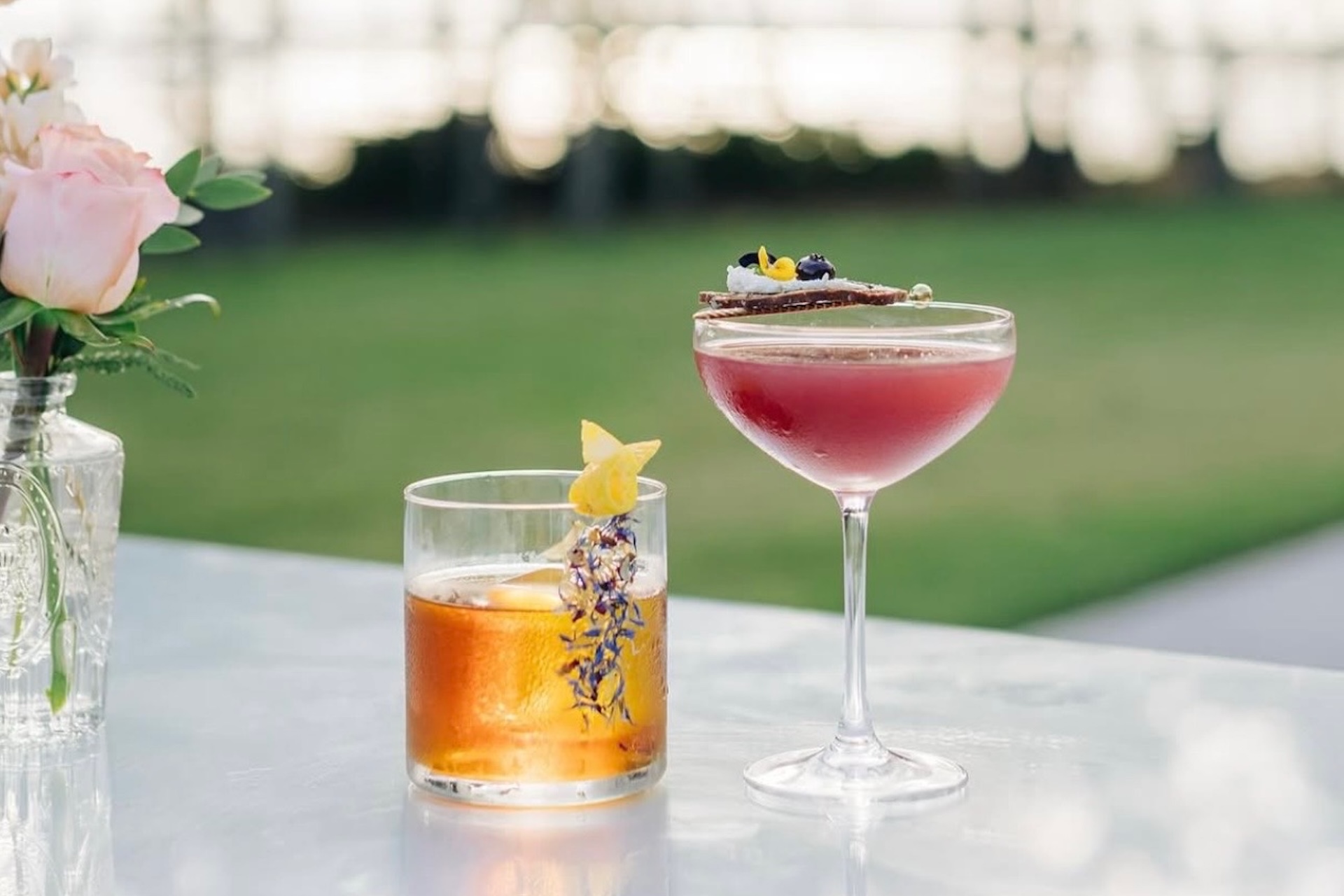 A glass of amber cocktail with a lemon twist and lavender sprigs sits beside a coupe glass with a reddish-pink drink garnished with chocolate and flowers on an outdoor table, with a vase of pink roses nearby.
