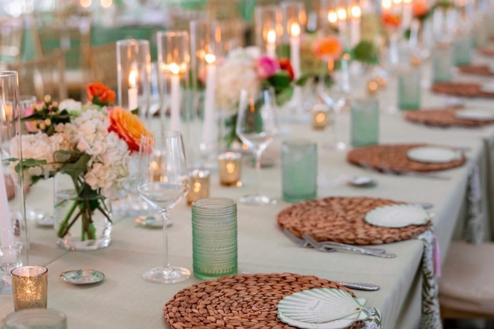 A long, elegant table set for a formal event with woven placemats, glassware, green-tinted glasses, candles in glass holders, and floral centerpieces with pink, orange, and white flowers.