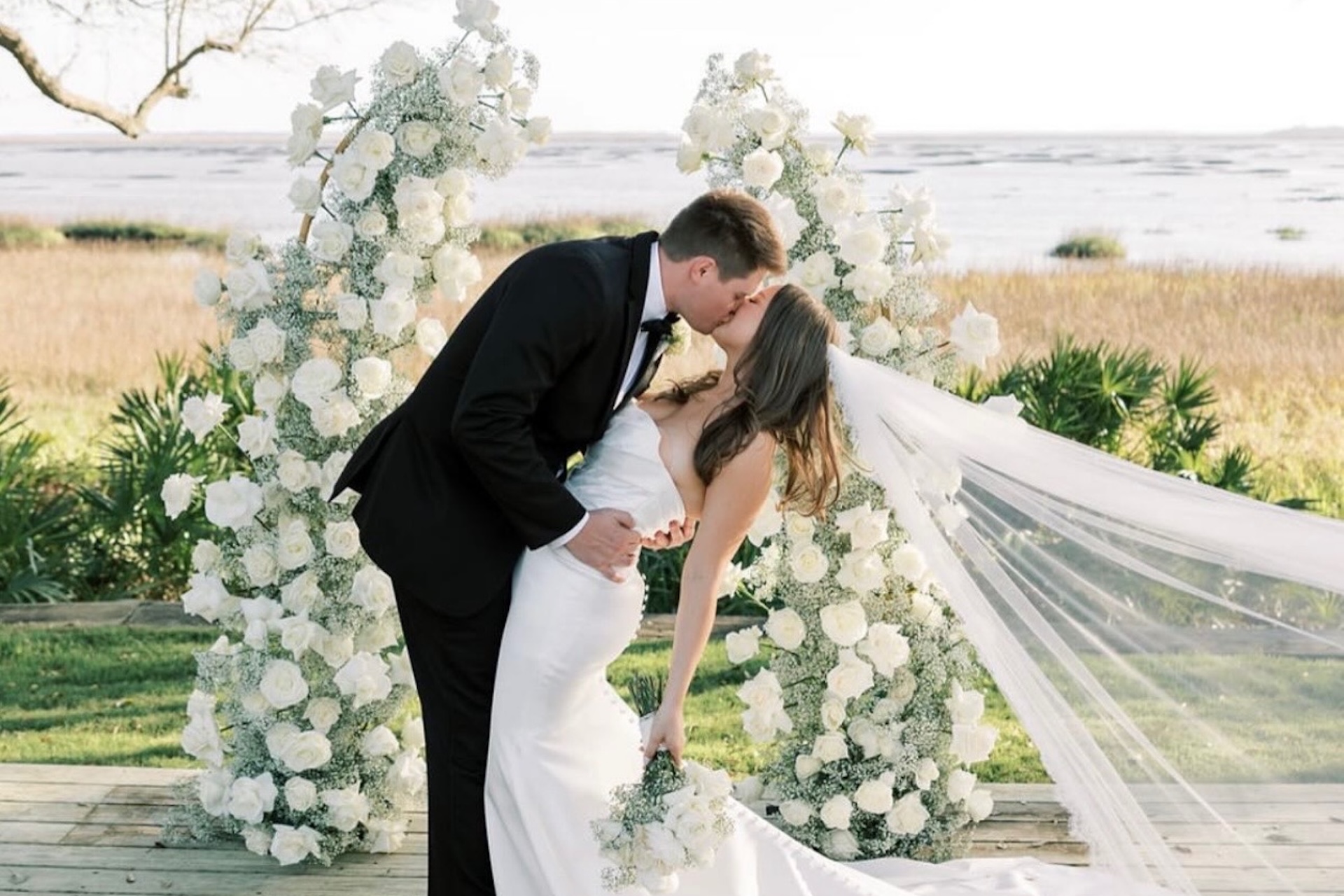 A bride and groom kiss at an outdoor wedding ceremony, surrounded by white flowers and greenery, with water and fields in the background. The bride’s veil flows behind her as the groom dips her.