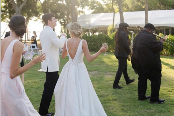 A bride and groom, both in white, walk outdoors with a bridesmaid holding drinks. Two musicians, one with a trombone, lead the group across a grassy area near white tents and trees.