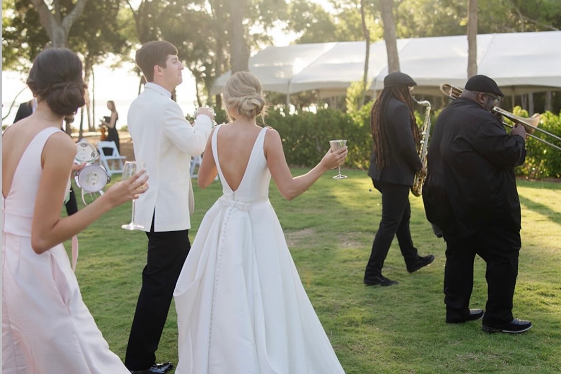 A bride and groom, both in white, walk outdoors with a bridesmaid holding drinks. Two musicians, one with a trombone, lead the group across a grassy area near white tents and trees.