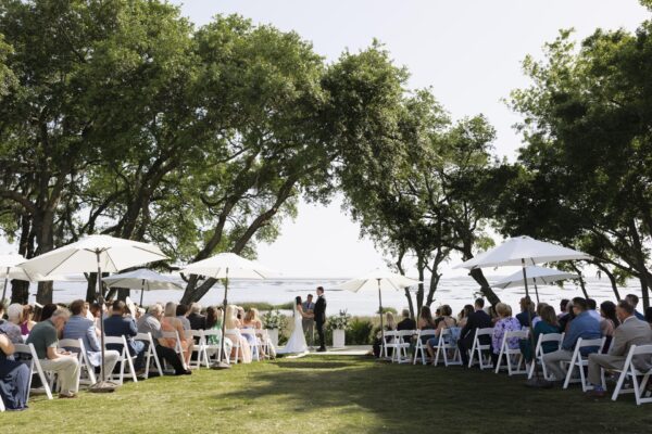 An outdoor wedding ceremony with a couple standing at the altar under trees, surrounded by seated guests on white chairs and white umbrellas, with a scenic view of water and greenery in the background.