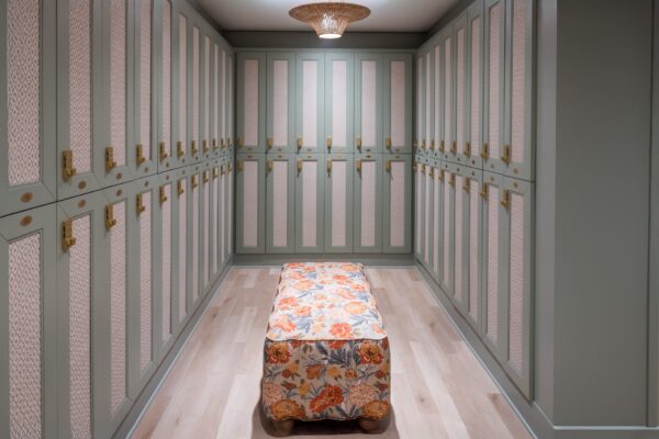 A spacious walk-in closet with light green cabinets, gold handles, and a floral-patterned ottoman in the center. The floor is light wood, and a woven ceiling light hangs above.