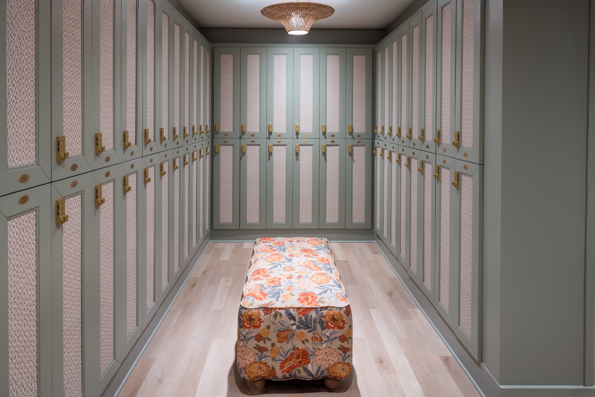 A spacious walk-in closet with light green cabinets, gold handles, and a floral-patterned ottoman in the center. The floor is light wood, and a woven ceiling light hangs above.