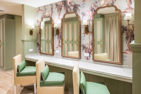Elegant dressing room with three vanity mirrors, green-accented chairs, marble countertop, and wall sconces. Floral wallpaper and paneled cabinets with cane doors add a stylish, vintage touch. Neutral and green tones throughout.