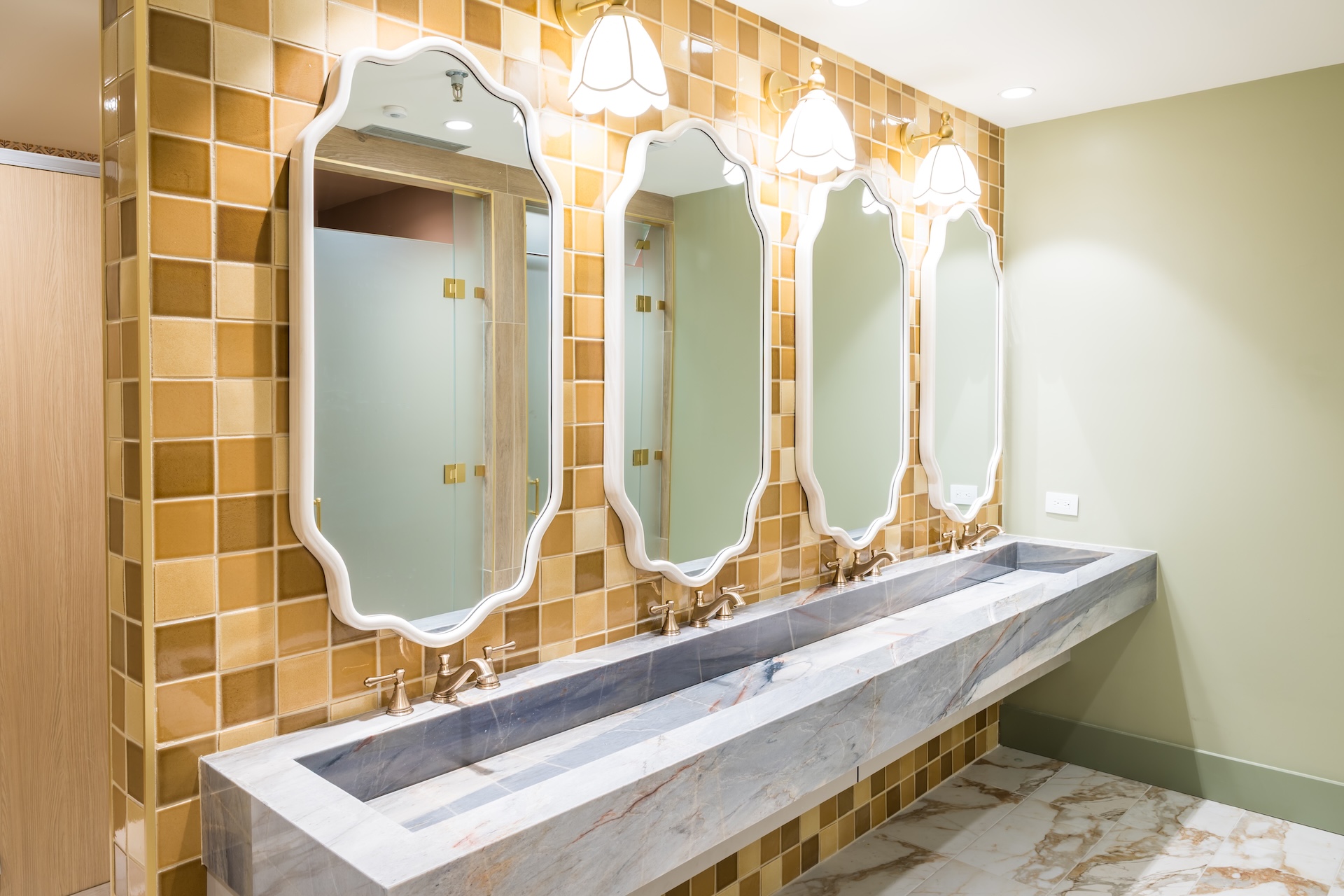 A modern restroom with three ornate mirrors above a long gray marble sink, gold faucets, yellow-brown tiled walls, and pendant lights. The floor is marble-patterned, and part of a stall is visible to the left.