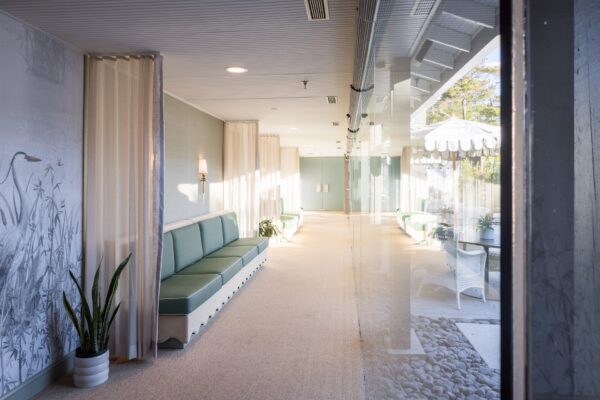 A bright, airy hallway with green cushioned seating, light curtains, a potted plant, and a large glass window reflecting the space and revealing an outdoor patio area with wicker chairs and a white scalloped awning.