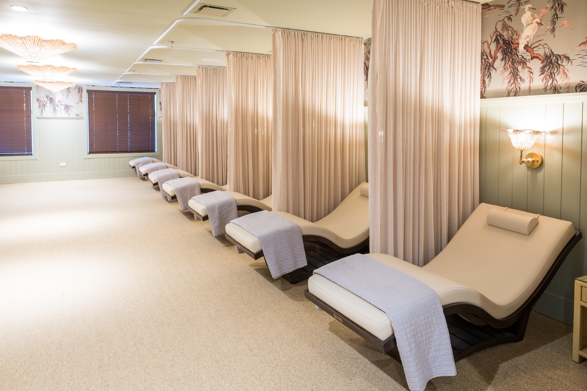 A serene spa relaxation room with several white recliner chairs, each separated by beige curtains, and topped with rolled pillows and folded light blue blankets. Soft lighting and elegant décor create a calm atmosphere.
