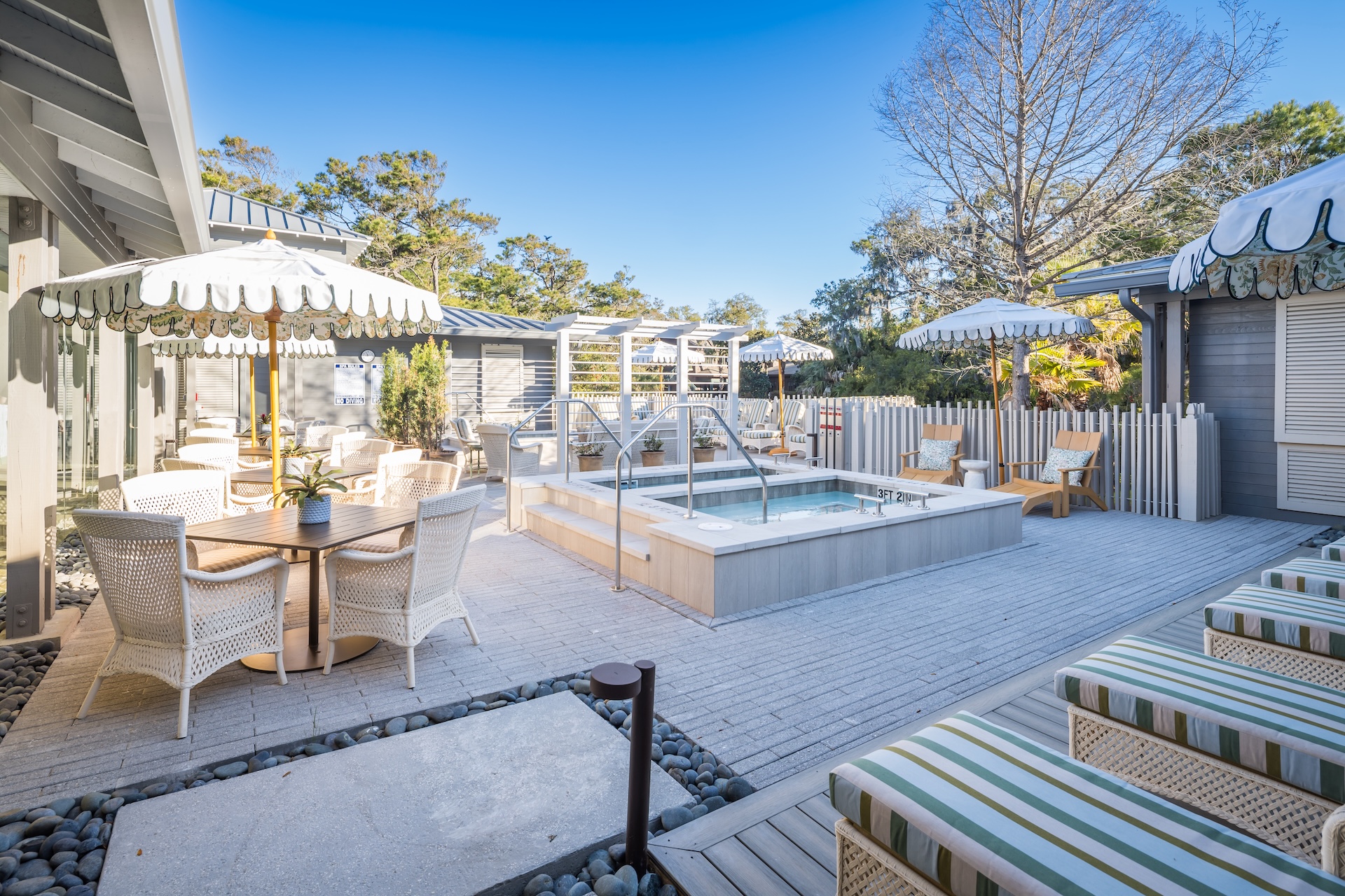 A sunny outdoor patio features a small pool or hot tub, striped lounge chairs, round tables with wicker chairs, and white umbrellas with fringe, surrounded by greenery and trees.