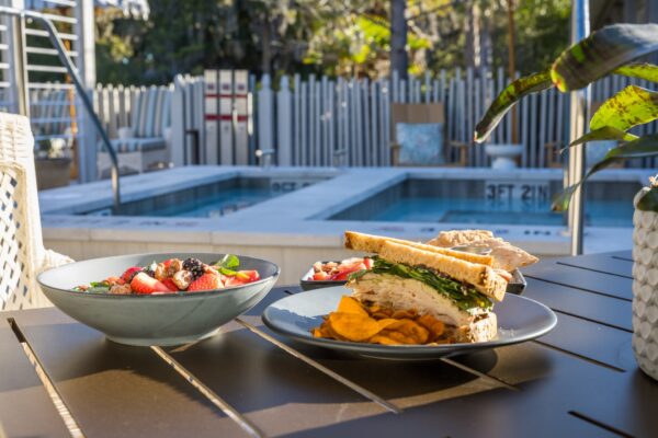 A turkey sandwich with chips, a salad with strawberries and blackberries, and another bowl of salad are set on a table beside a swimming pool in a sunny outdoor setting.
