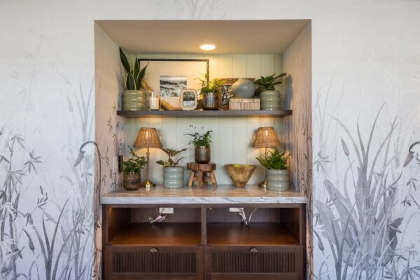 A built-in wall nook with two wooden shelves displaying green pottery, plants, framed art, and table lamps, set above a marble countertop and dark wood drawers. The background features pale botanical wallpaper.