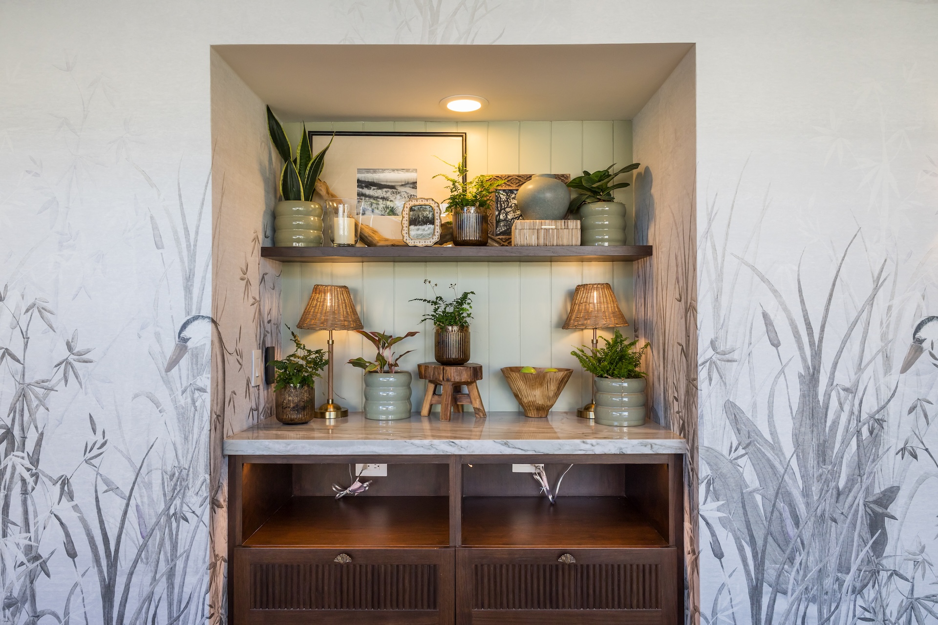 A built-in wall nook with two wooden shelves displaying green pottery, plants, framed art, and table lamps, set above a marble countertop and dark wood drawers. The background features pale botanical wallpaper.