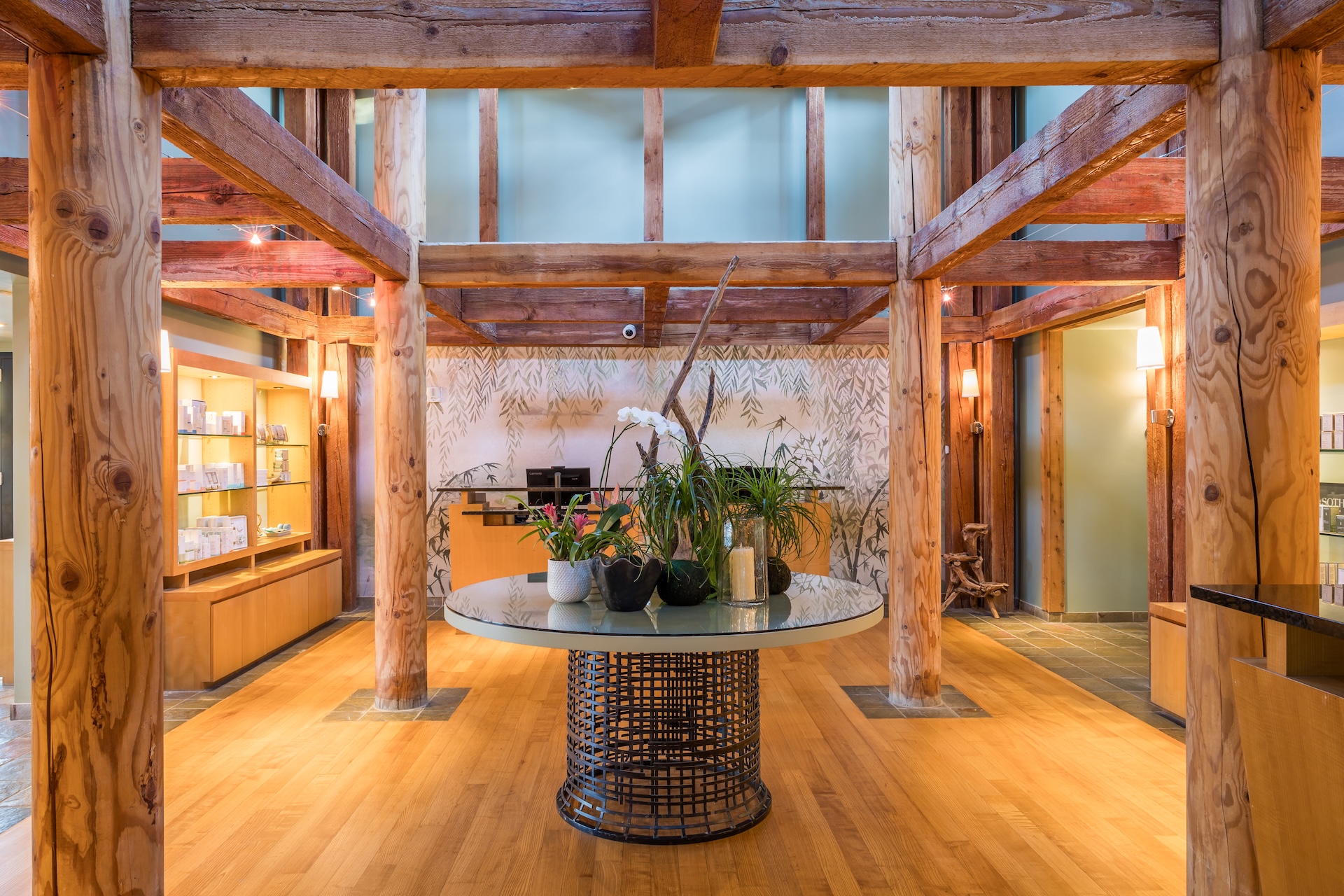 A spacious, rustic lobby with wooden beams and posts, featuring a round table with potted plants and orchids in the center on a wooden floor, with shelves and reception desk in the background.
