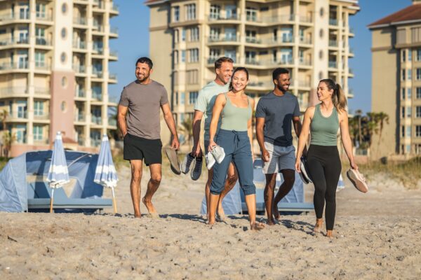 Five people walk barefoot on a sandy beach, smiling and holding their shoes, with beach tents, umbrellas, and tall apartment buildings in the background under a clear blue sky.