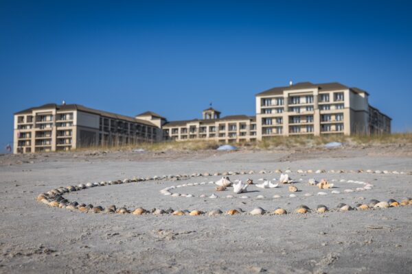 A sandy beach with a large circular arrangement of seashells in the foreground and several multi-story beachfront hotels in the background under a clear blue sky.