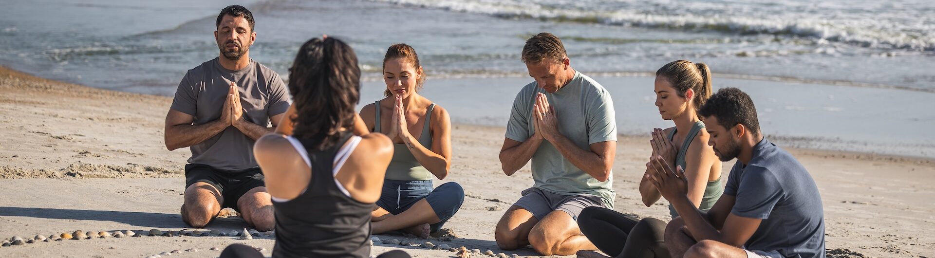 A group of people sit cross-legged in a circle on the beach, eyes closed and hands in prayer position, meditating together near the ocean. A woman faces the group, leading the session.