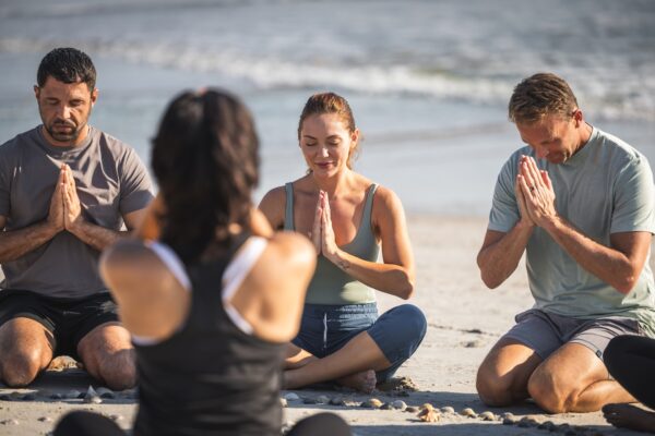 Three adults sit cross-legged on the beach with eyes closed, hands pressed together in prayer pose, and a yoga instructor sits facing them, guiding a meditation session by the ocean.