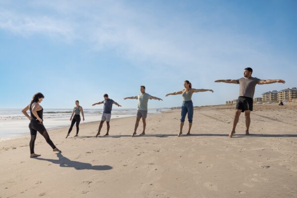 A group of six people, including an instructor, practice stretching exercises on a sandy beach. They are spaced out in a line, arms extended, with buildings and blue sky in the background.