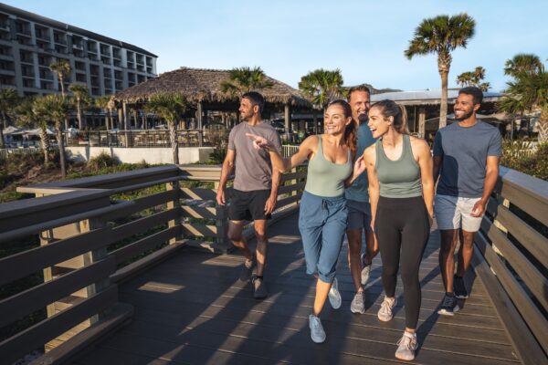 Five adults walk together on a wooden boardwalk near palm trees and a thatched-roof building, smiling and enjoying a sunny day. One woman in front is pointing ahead, and everyone appears relaxed and happy.