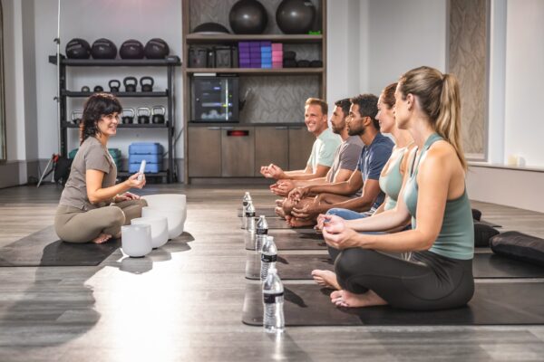 A yoga instructor sits cross-legged facing a group of five people seated on mats in a gym studio, all meditating. Yoga blocks, kettlebells, and water bottles are visible in the background.