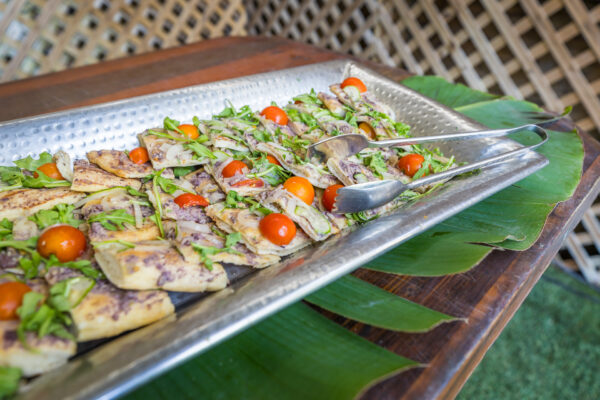 A metal tray filled with sliced flatbread topped with arugula, cherry tomatoes, and other ingredients sits on green leaves, with serving tongs placed on the tray.