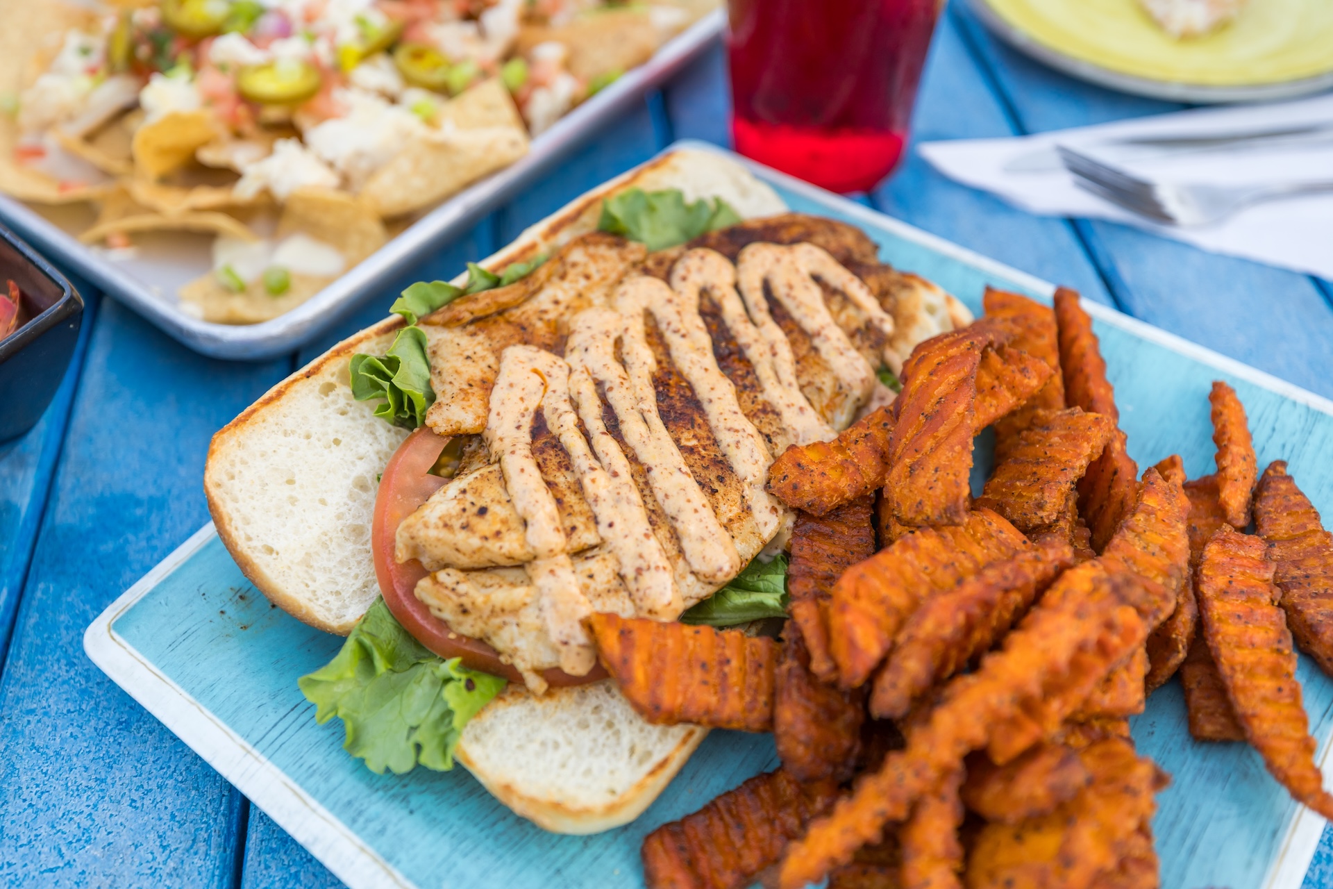 A grilled chicken sandwich with lettuce and tomato, drizzled with sauce, served with a side of crinkle-cut sweet potato fries on a blue plate. In the background, nachos and a red drink are visible.