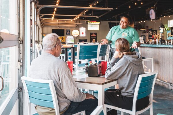 A server in a green shirt smiles and talks to two seated customers at a casual restaurant with teal chairs and large windows. The customers look engaged and the table has drinks and condiments.