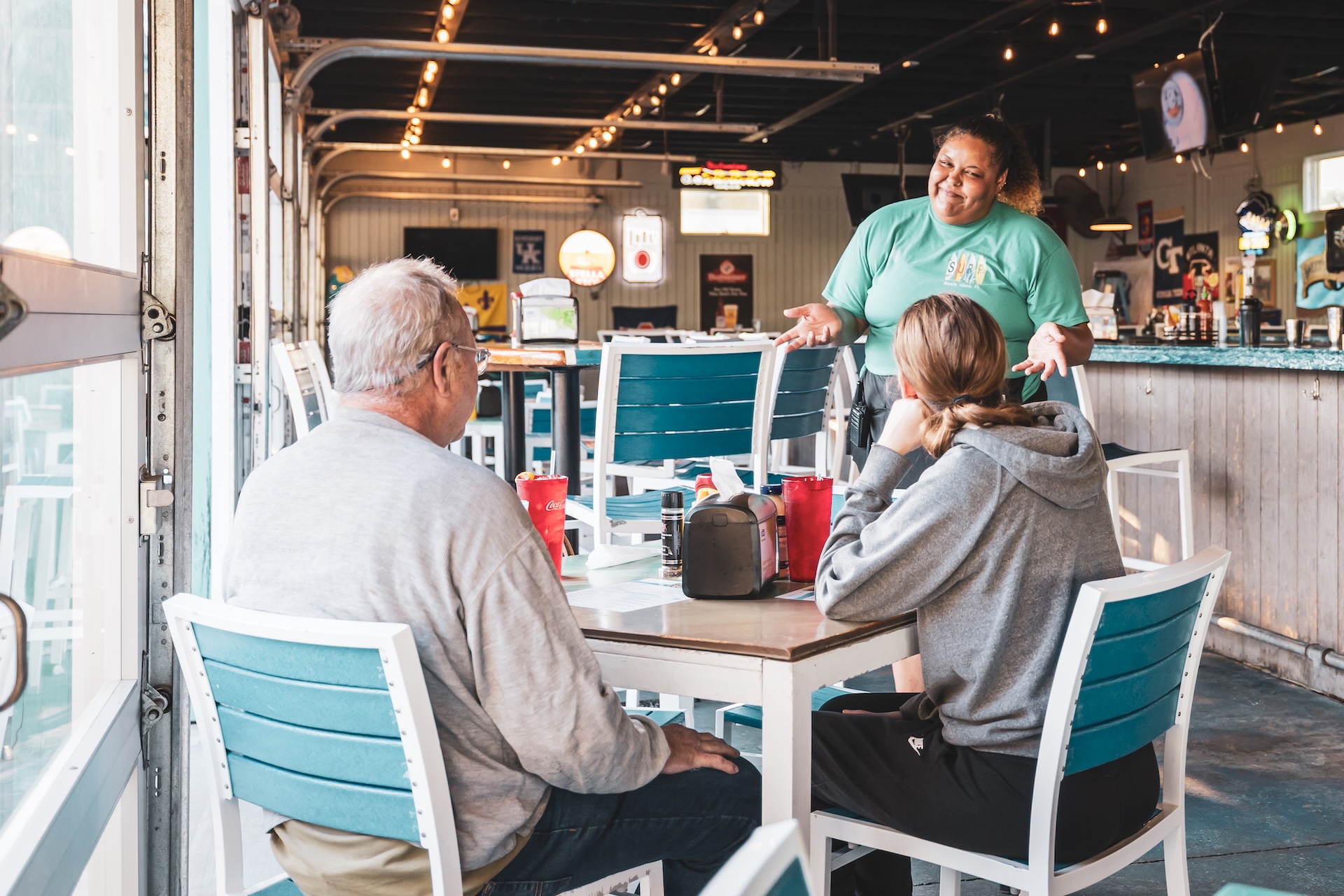 A server in a green shirt smiles and talks to two seated customers at a casual restaurant with teal chairs and large windows. The customers look engaged and the table has drinks and condiments.