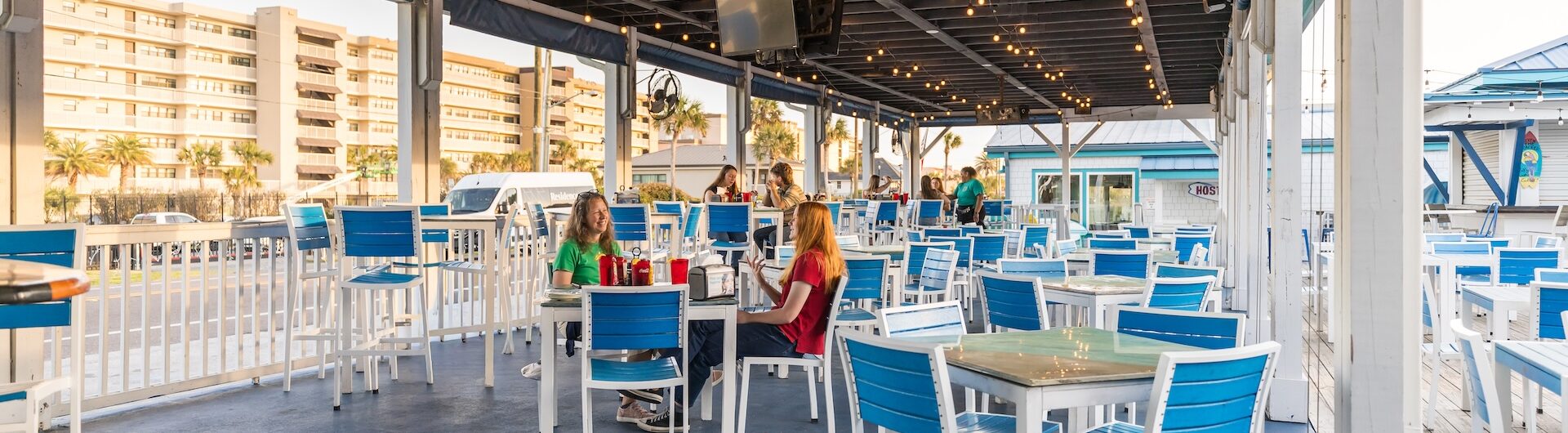 Outdoor restaurant patio with blue and white chairs and tables, a few people sitting and talking, string lights overhead, and buildings visible in the background on a sunny day.