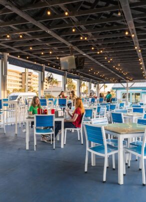 Outdoor restaurant patio with blue and white chairs and tables, a few people sitting and talking, string lights overhead, and buildings visible in the background on a sunny day.