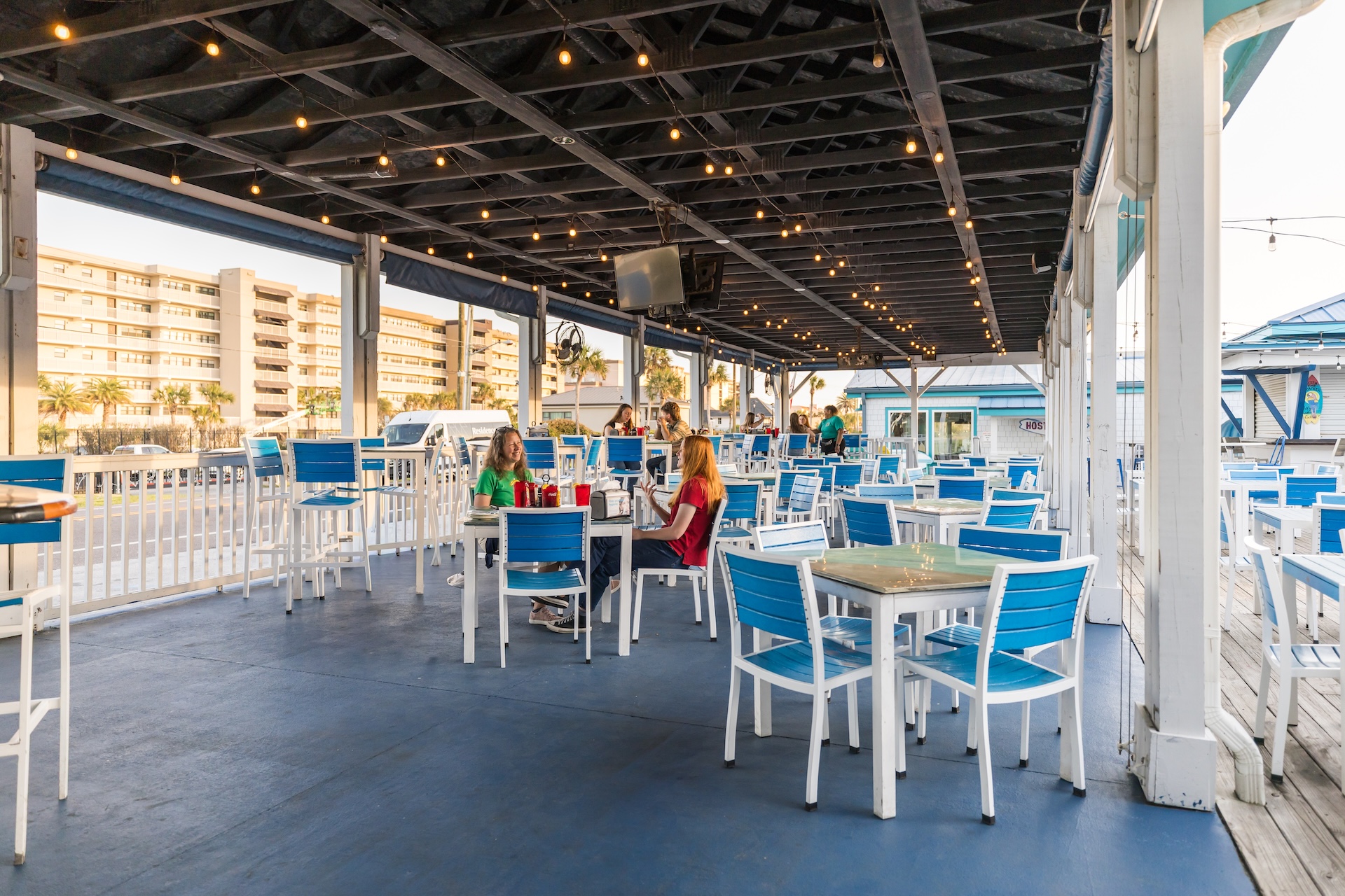 Outdoor restaurant patio with blue and white chairs and tables, a few people sitting and talking, string lights overhead, and buildings visible in the background on a sunny day.