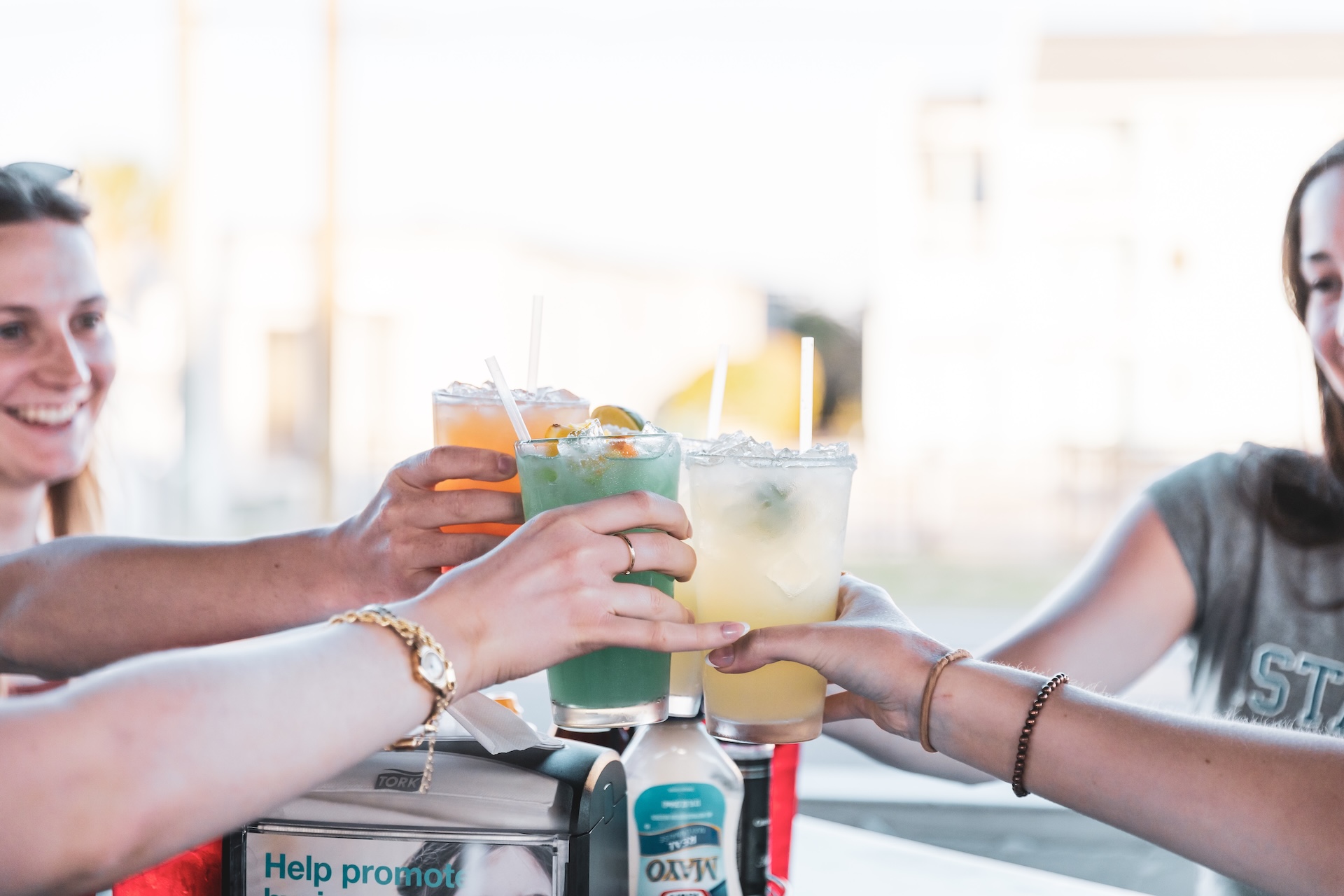 Four people smile and raise colorful drinks in a toast at an outdoor table, enjoying a sunny day together. Bottles and napkins are visible on the table in the foreground.