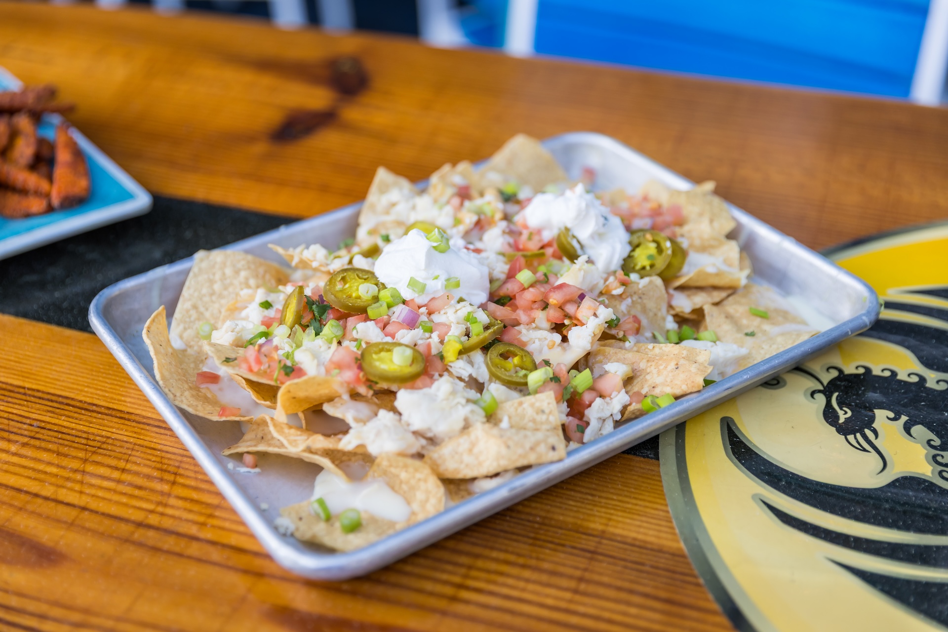 A tray of nachos topped with diced tomatoes, jalapeños, green onions, melted cheese, and dollops of sour cream sits on a wooden table with a dragon design partially visible.