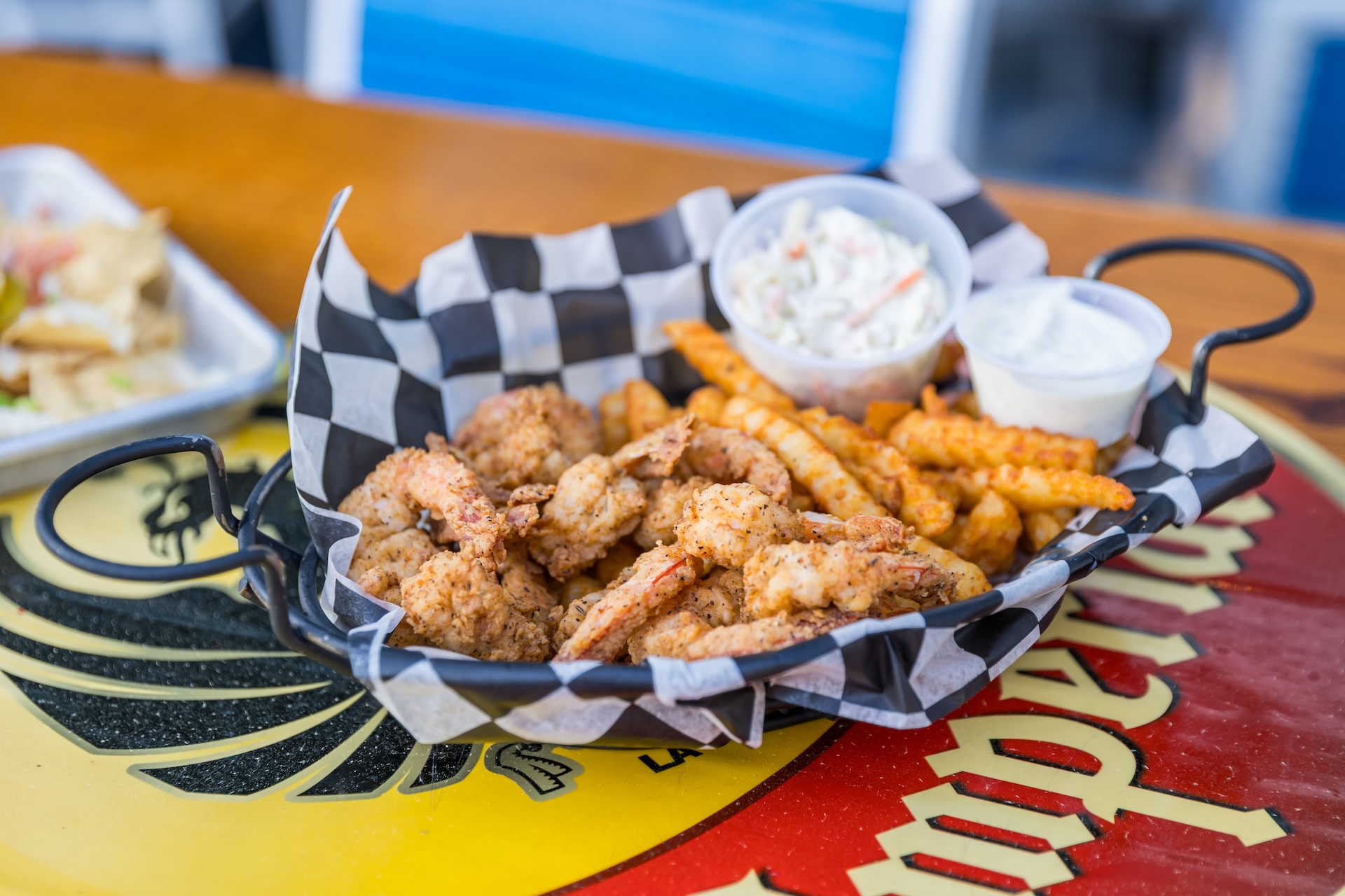 A basket lined with checkered paper contains fried shrimp, crinkle-cut fries, a cup of coleslaw, and a cup of dipping sauce, set on a colorful tabletop.