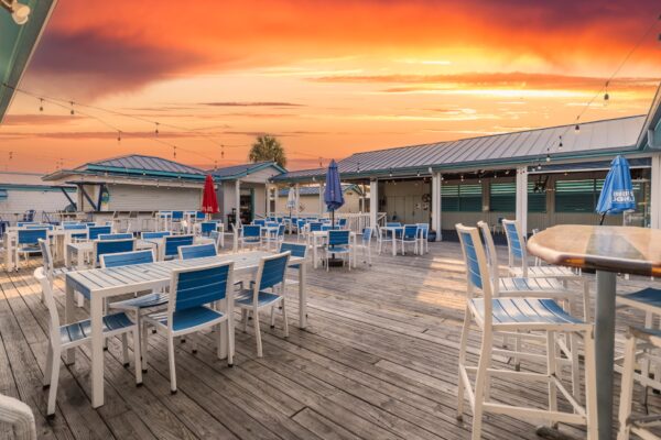 Outdoor restaurant patio with blue and white tables and chairs on a wooden deck, string lights overhead, umbrellas closed, and a vivid orange sunset sky in the background. The area appears empty and peaceful.