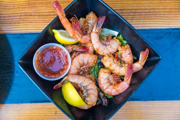 A black square bowl filled with seasoned cooked shrimp, lemon wedges, leafy greens, and a small container of red dipping sauce, placed on a wooden table.