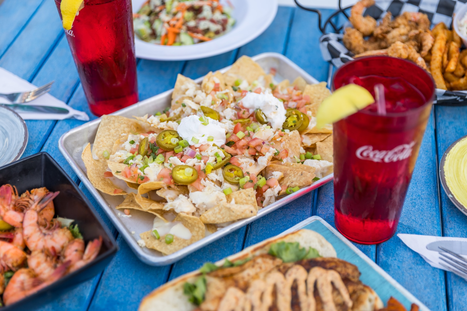 A tray of nachos topped with jalapeños, sour cream, diced tomatoes, and green onions sits on a blue table alongside plates of shrimp, fried food, and two red drinks with lemon wedges.