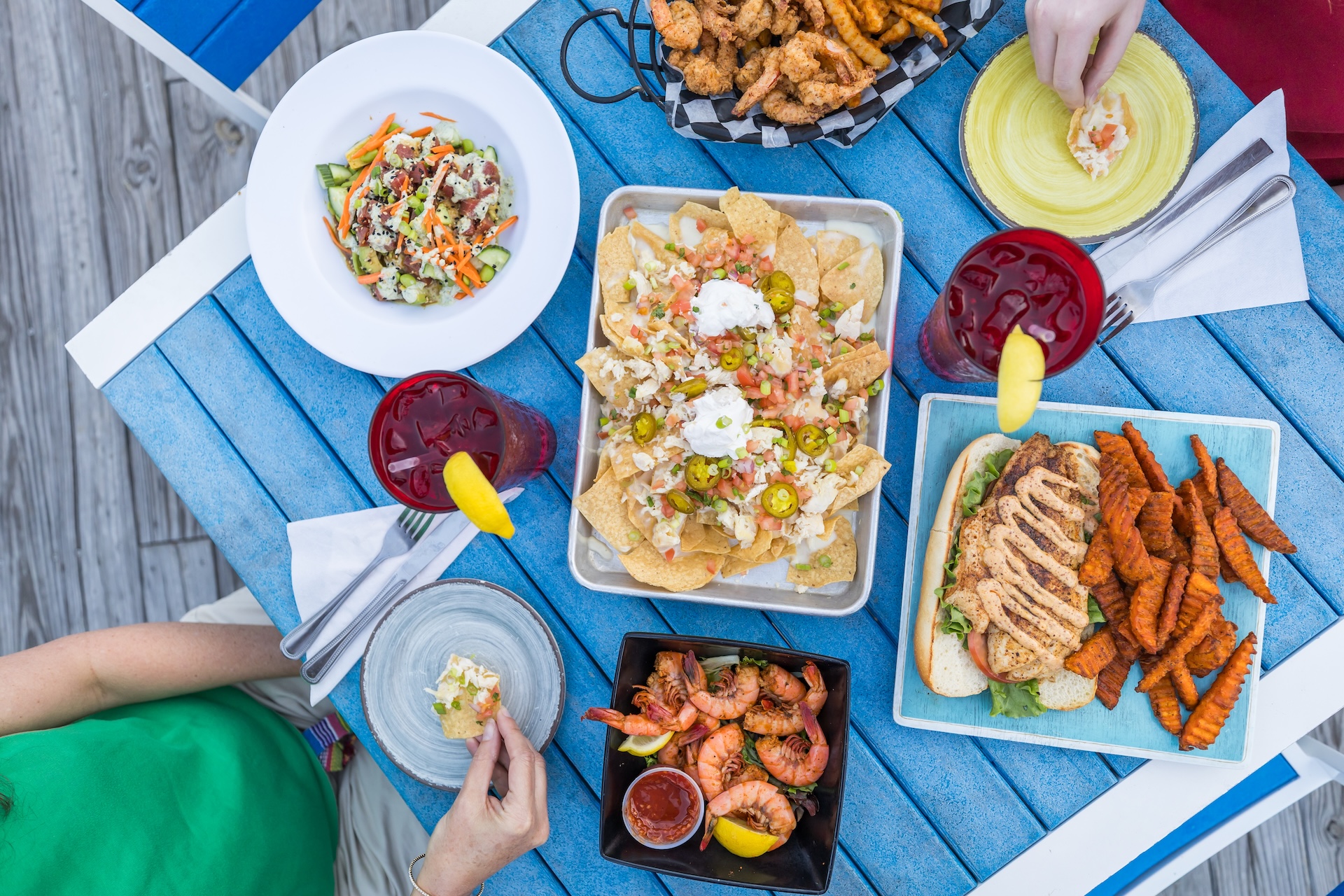 A table set with nachos, grilled chicken and sweet potato fries, shrimp, fried seafood, salads, and iced drinks with lemon, viewed from above with people reaching for food.