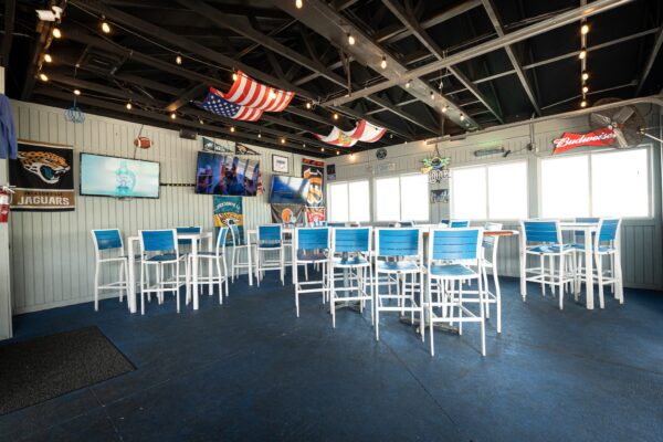 A bright bar with blue and white chairs and tables, sports memorabilia on the walls, TVs, hanging lights, and American flags decorating the ceiling. Sunlight streams in through large windows.
