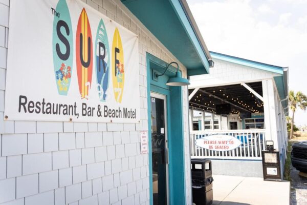 A beachside building with a sign reading The Surf Restaurant, Bar & Beach Motel featuring colorful surfboards. Nearby, another sign says Please wait to be seated outside a covered patio with string lights.