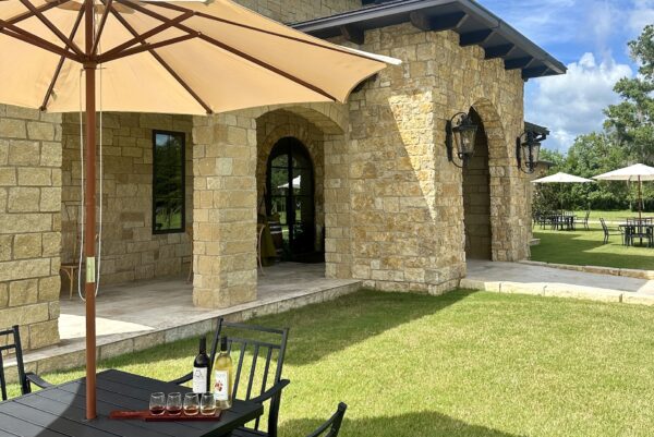 A sunlit patio at a stone building, with black metal tables and chairs on green grass. A wine bottle and glasses sit on a table under a large umbrella. Other tables and umbrellas are spread out in the background.