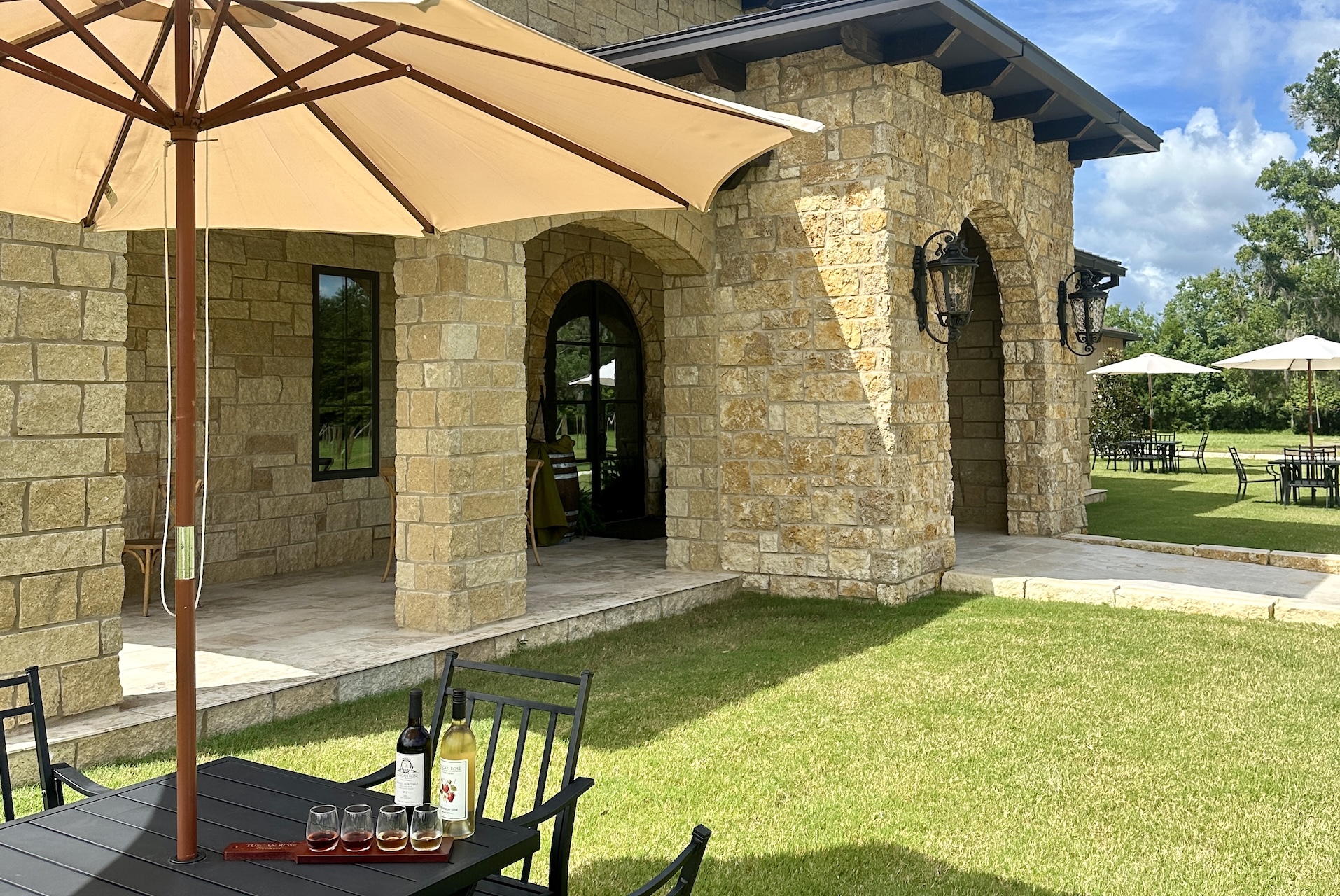 A sunlit patio at a stone building, with black metal tables and chairs on green grass. A wine bottle and glasses sit on a table under a large umbrella. Other tables and umbrellas are spread out in the background.