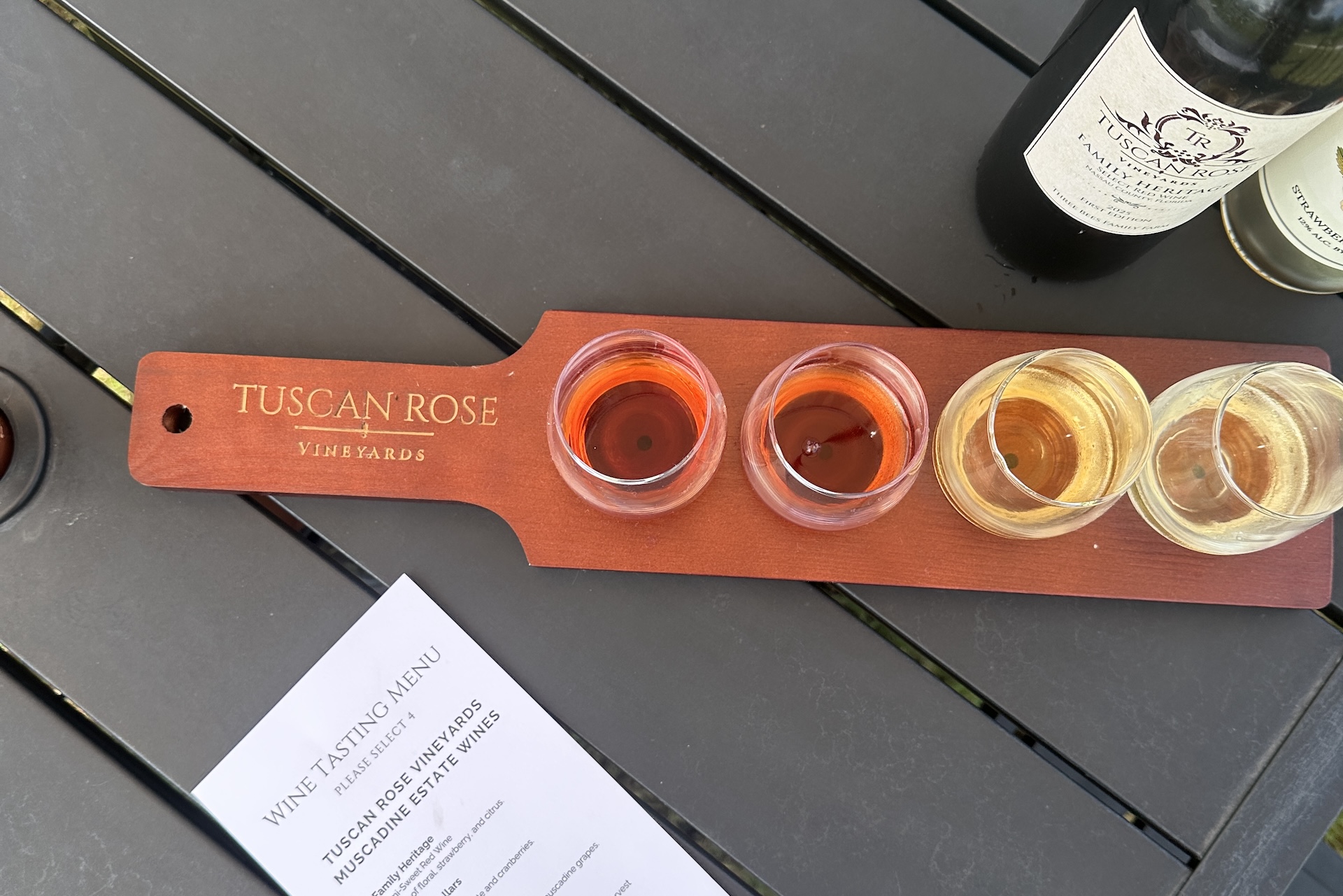 A wooden flight board labeled Tuscan Rose Vineyards holds four glasses of wine, ranging in color from red to white, on a dark table next to wine bottles and a printed wine tasting menu.
