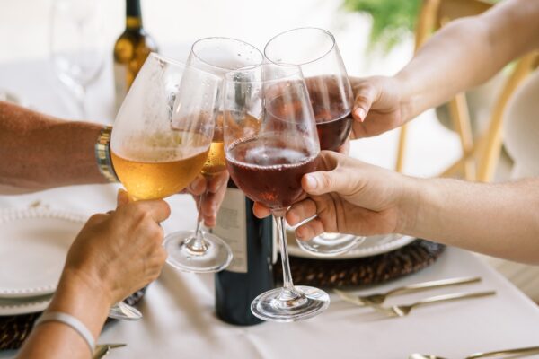 Four people clinking wine glasses filled with red wine and a golden drink over a white table set with plates, cutlery, and a bottle of wine, celebrating or toasting together.