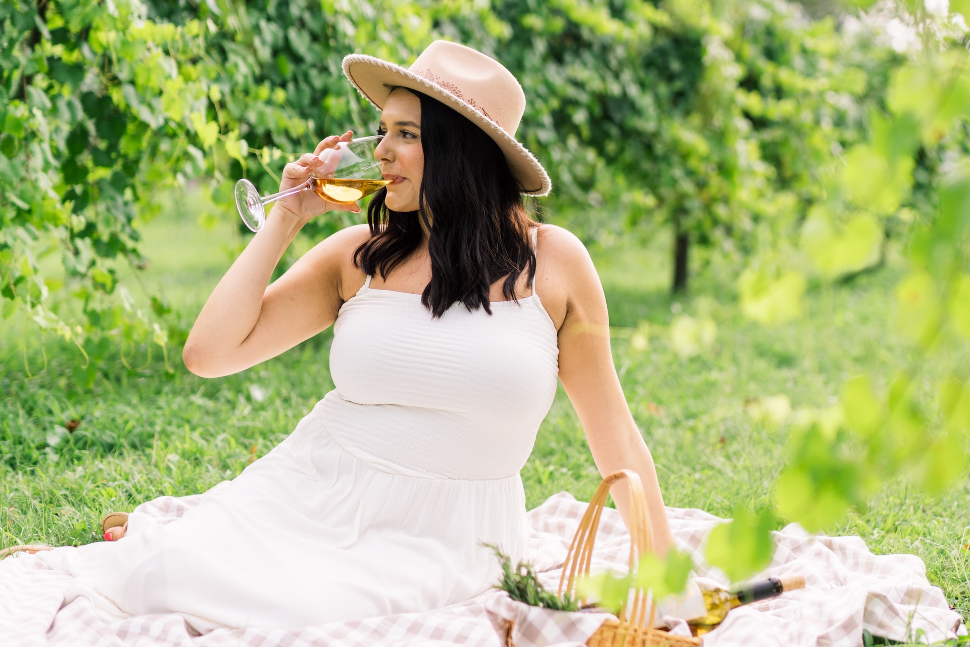 A woman in a white dress and hat sits on a picnic blanket outdoors, sipping wine from a glass. She is surrounded by greenery, with a picnic basket beside her on the grass.